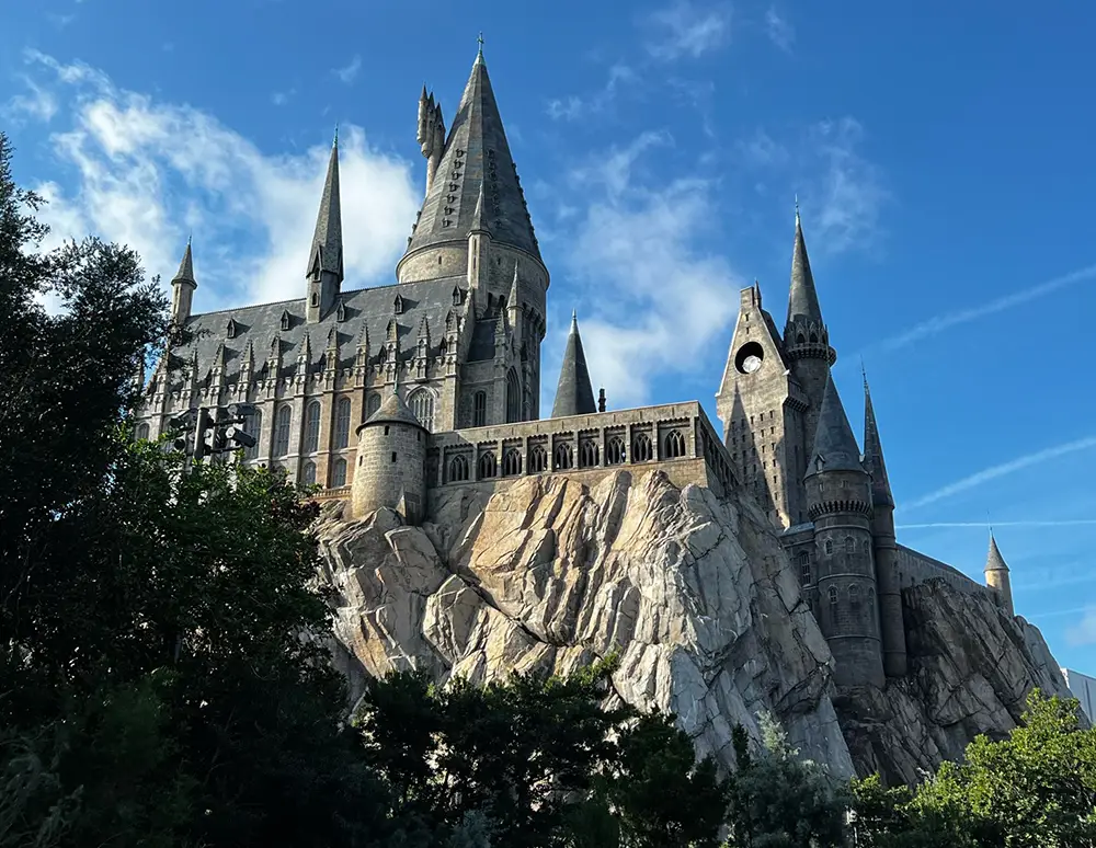 a Large, Stone Castle with Multiple Tall Spires and Towers Sits Atop a Rocky Cliff at Universal Orlando Resort, Surrounded by Green Trees Under a Clear Blue Sky. - Favorite Grampy Travels - Favorite Grampy Travels 2026