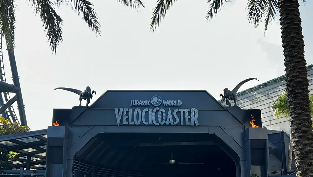 Entrance to the Jurassic World Velocicoaster Ride at Universal Orlando Resort, Featuring Two Raptor Statues on the Roof and Surrounded by Palm Trees Under a Bright Sky. - Favorite Grampy Travels - Favorite Grampy Travels 2026 - Favorite Grampy Travels 2026