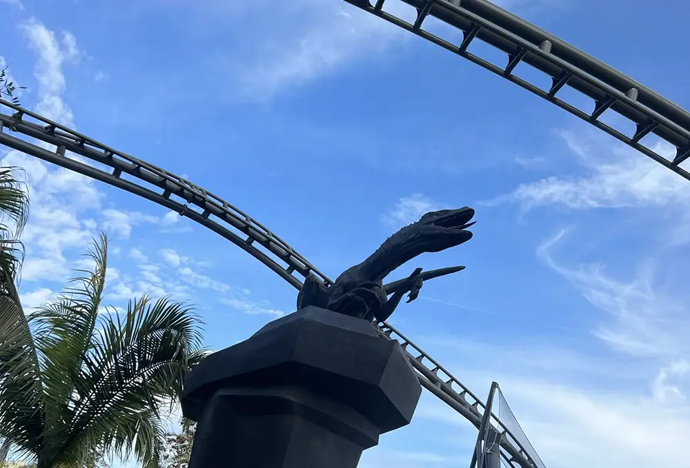 a Statue of a Velociraptor on a Pedestal is Shown Beneath the Looping Tracks of Jurassic World Velocicoaster at Universal Orlando Resort, with Palm Trees and a Blue Sky in the Background. - Favorite Grampy Travels - Favorite Grampy Travels 2026