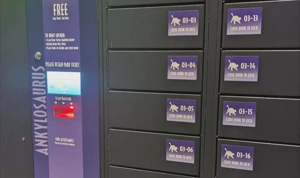 a Row of Black Lockers with Purple Labels Numbered 03-04 to 03-16 Sits Beside an Ankylosaurus Sign Featuring Instructions and a Scanner for Free Locker Use at Universal Orlando’s Epic Universe. - Favorite Grampy Travels - Favorite Grampy Travels 2026