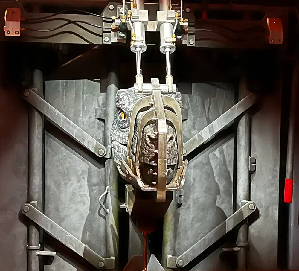 a Mechanical Dinosaur Head, Possibly a Raptor from Universal’s Jurassic World Velocicoaster, is Secured by Metal Clamps and Rods Against a Dark, Industrial Background, with Yellow-lit Eyes Peering Through the Restraints. - Favorite Grampy Travels - Favorite Grampy Travels 2026 - Favorite Grampy Travels 2026