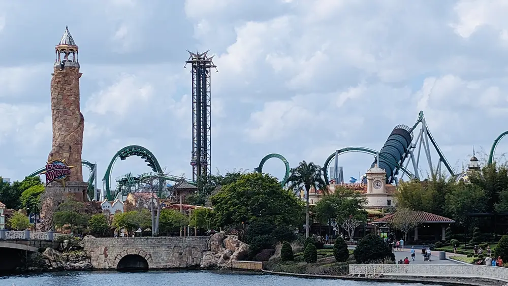 a Theme Park Scene with a Tall Stone Tower, Roller Coasters with Green Tracks Like Hagrid - Favorite Grampy Travels 2026's Magical Creature Motorbike Adventure, and various buildings surrounded by trees next to a body of water under a cloudy sky. - Favorite Grampy Travels