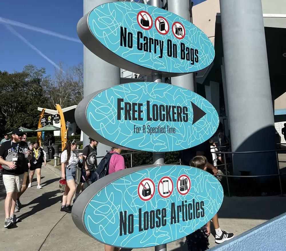 Uor-universal-studios-men-in-black-lockers-sign - Favorite Grampy Travels Three Blue Signs at Universal Orlando Read No Carry on Bags, Free Lockers for a Specified Time, and No Loose Articles. People Walk Around the Theme Park in the Background on a Sunny Day. - Favorite Grampy Travels - Favorite Grampy Travels 2026