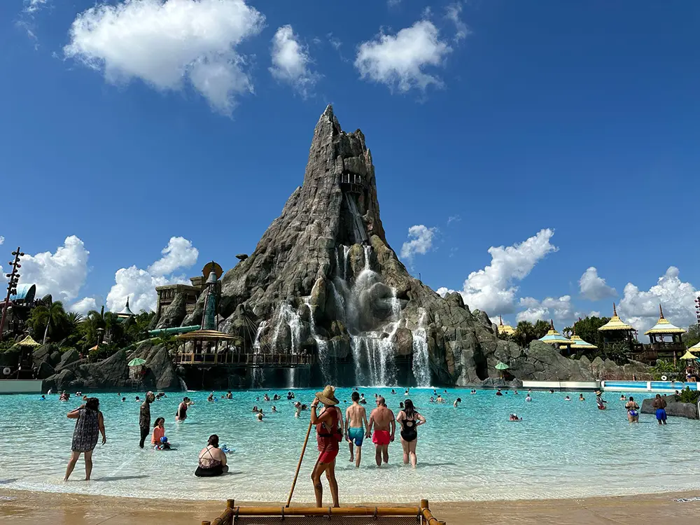 People Swim and Relax in a Large Wave Pool with a Tall, Rocky Volcano and Waterfalls at Universal Orlando Resort’s Water Park, All Beneath a Bright Blue Sky Dotted with Scattered Clouds. - Favorite Grampy Travels - Favorite Grampy Travels 2026