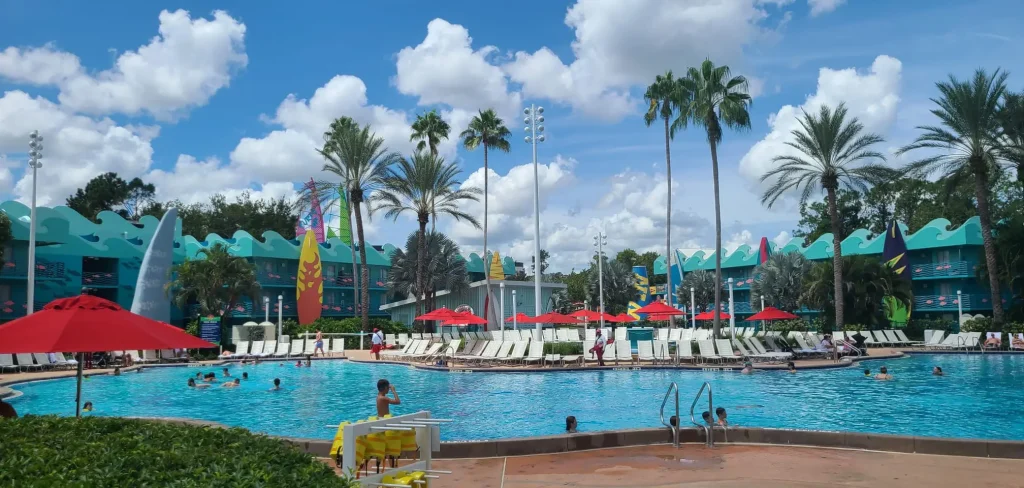 a Large Outdoor Swimming Pool at a Walt Disney World Resort, Surrounded by Lounge Chairs, Red Umbrellas, Palm Trees, and Colorful Buildings on a Sunny Day with Blue Sky and Fluffy Clouds. Guests Are Swimming and Relaxing by the Pool. - Favorite Grampy Travels - Favorite Grampy Travels 2026