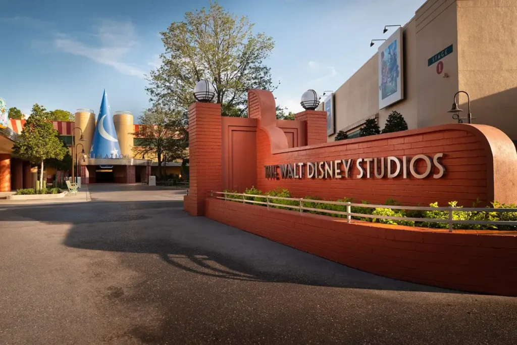 Entrance to the Walt Disney Studios with a Red Brick Wall Featuring the Studio Name in Large Letters, Trees, Lampposts, and a Blue Sorcerer’s Hat Structure in the Background Under a Clear Sky. the Location is the Animation Courtyard at Disney - Favorite Grampy Travels 2026's Hollywood Studios in Walt Disney World - Favorite Grampy Travels