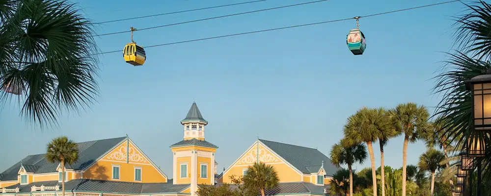 Two Colorful Gondolas Glide Above a Yellow Building with a Blue Roof and Cupola at Disney World - Favorite Grampy Travels 2026's Caribbean Beach, surrounded by palm trees under a clear blue sky. - Favorite Grampy Travels