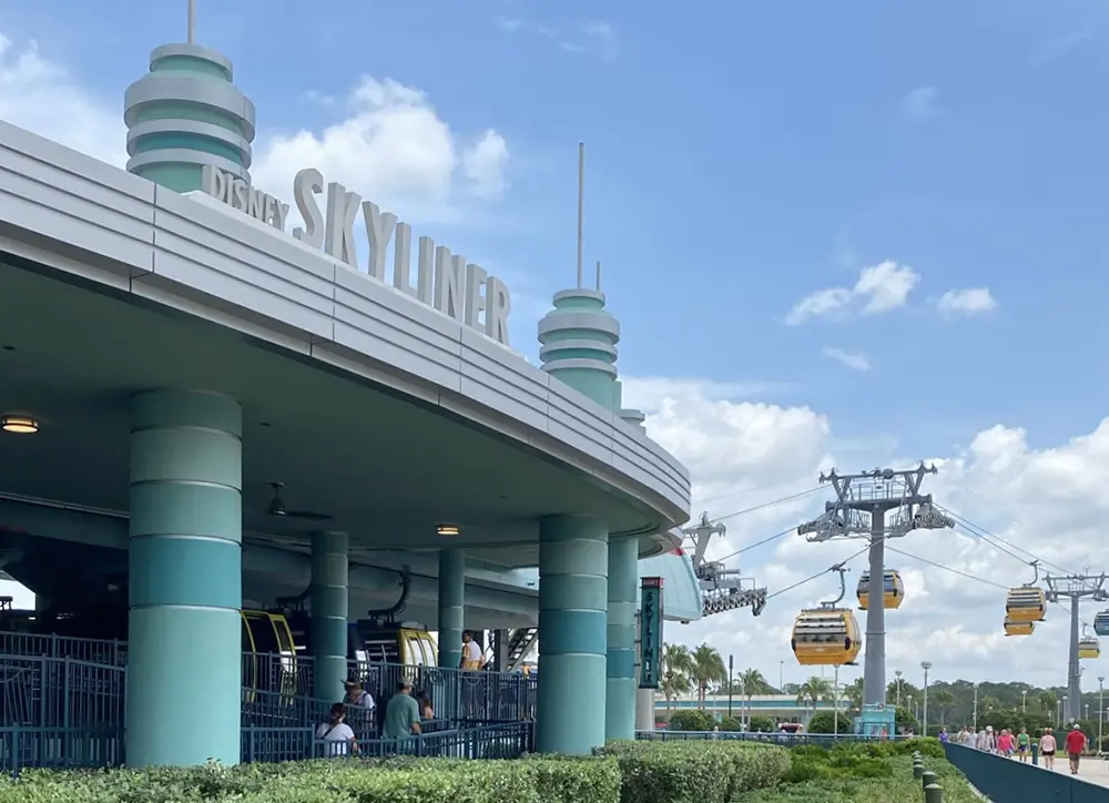 a Covered Station Marked Disney Skyliner with People Waiting Underneath, and Several Colorful Gondolas Suspended on Cables in the Background Against a Blue Sky with Scattered Clouds. - Favorite Grampy Travels - Favorite Grampy Travels 2026