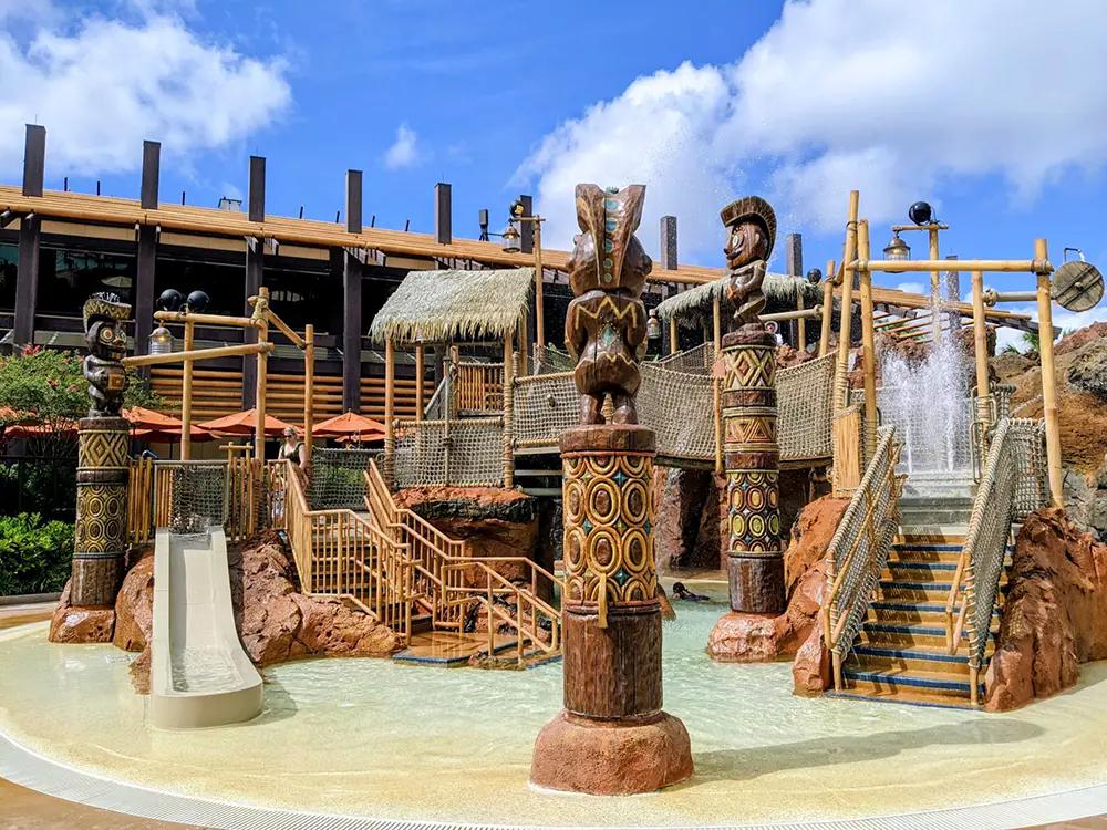 Wdw-polynesian-village-resort-kids-kiki-tikis-splash-play - Favorite Grampy Travels Children’s Splash Play Area with Polynesian-themed Wooden Statues, Water Sprays, and a Small Slide, Set Outdoors Near Disney - Favorite Grampy Travels 2026's Monorail Resorts with the Contemporary building and umbrellas visible in the background. - Favorite Grampy Travels