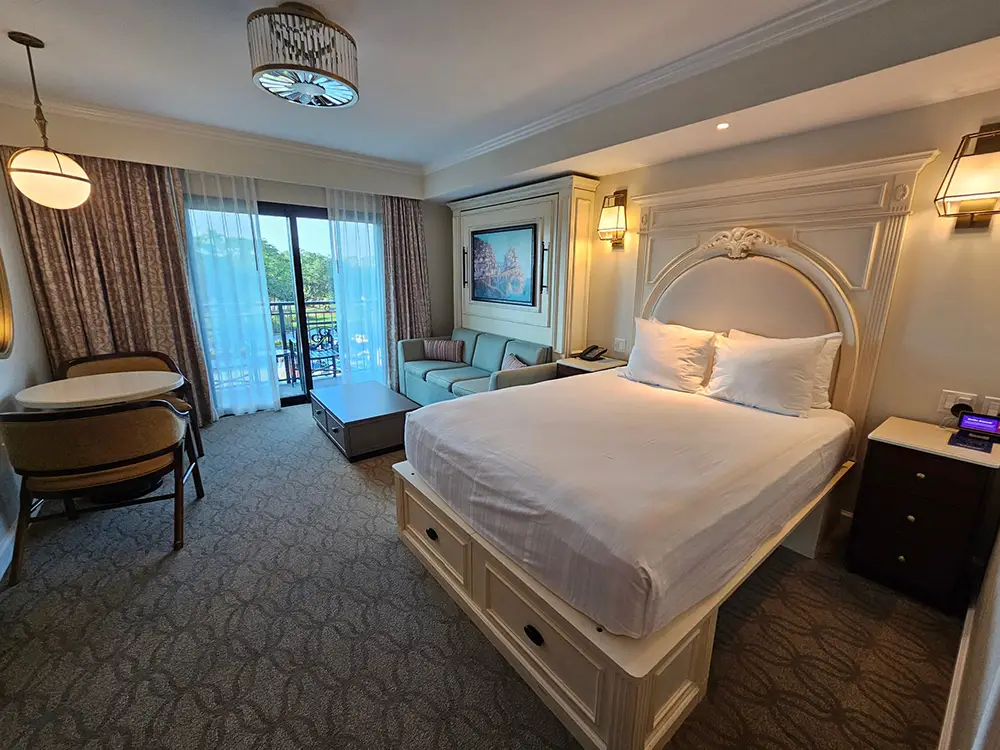 Wdw-riviera-resort-bedroom-interior - Favorite Grampy Travels a Hotel Room at Disney’s Riviera Resort with a Neatly Made Bed, Sofa, Small Round Table with Two Chairs, Coffee Table, Large Windows with Curtains, and a Balcony Overlooking Greenery Near the Convenient Skyliner Access. - Favorite Grampy Travels - Favorite Grampy Travels 2026