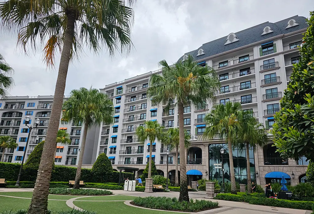 Wdw-riviera-resort-courtyard-exterior - Favorite Grampy Travels a Modern Multi-story Resort with Blue Accents, Surrounded by Palm Trees and Manicured Gardens at Walt Disney World. Under a Cloudy Sky, a Few People Walk Near the Entrance Featuring Blue Awnings. - Favorite Grampy Travels - Favorite Grampy Travels 2026