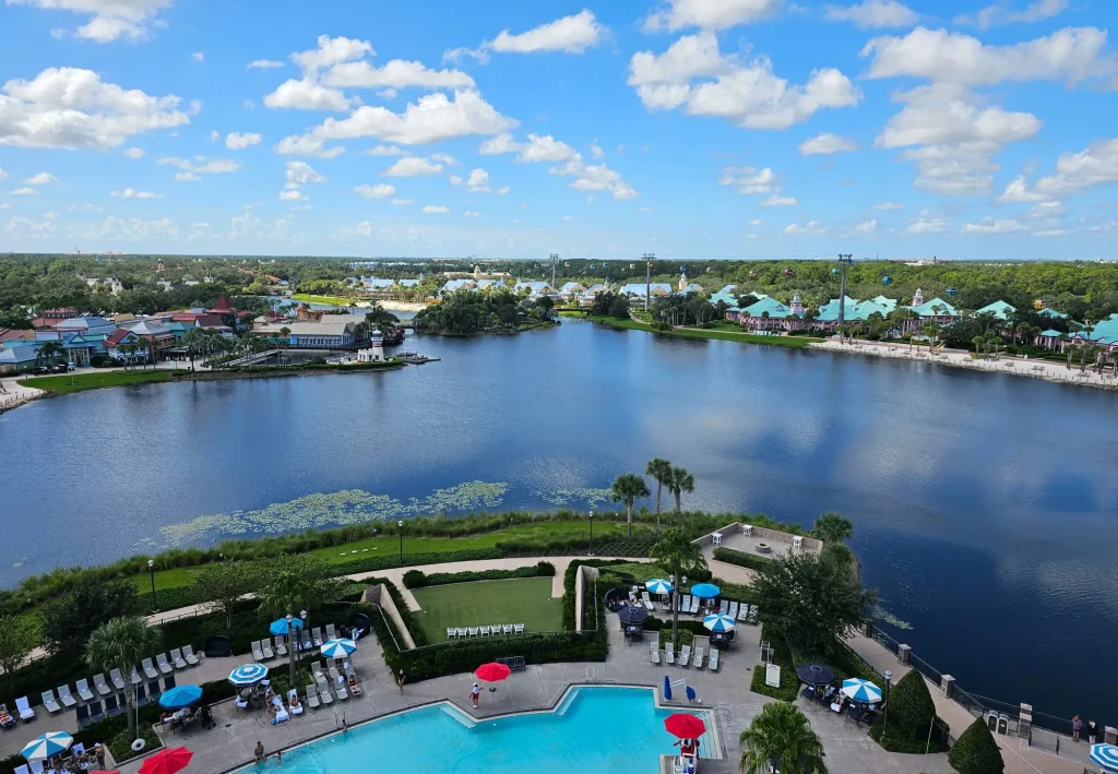 Wdw-riviera-resort-pool-courtyard-birds-eye-view-overlooking-lake-and-caribbean-beach - Favorite Grampy Travels a Scenic View of Disney - Favorite Grampy Travels 2026's Riviera Resort pool with lounge chairs and umbrellas in the foreground, a lake surrounded by greenery in the middle, and colorful buildings near the Walt Disney World Skyliner under a partly cloudy blue sky in the background. - Favorite Grampy Travels