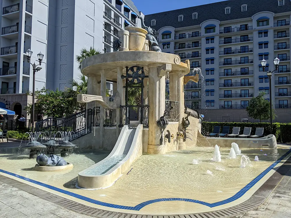 a Children’s Splash Pad with Small Slides, Fountains, and Water Features Shaped Like Animals and Vases Sits in Front of Disney - Favorite Grampy Travels 2026's Riviera Resort, a multi-story hotel with balconies. The sunny sky frames the nearby Skyliner in the background. - Favorite Grampy Travels
