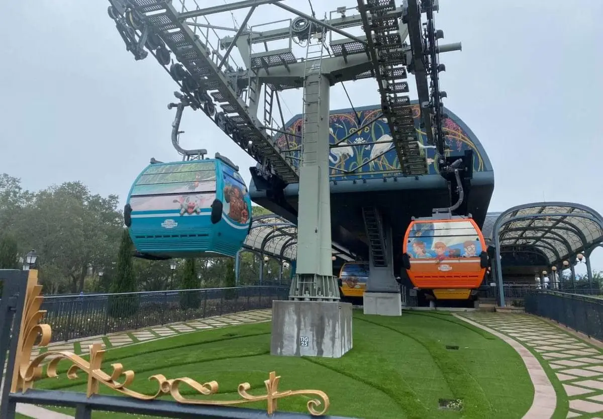 Two Colorful Gondola Cabins—one Blue and One Orange—of the Disney Skyliner Are Suspended from Cables at a Station with a Decorative Roof, Surrounded by Green Grass and a Black Fence. - Favorite Grampy Travels - Favorite Grampy Travels 2026
