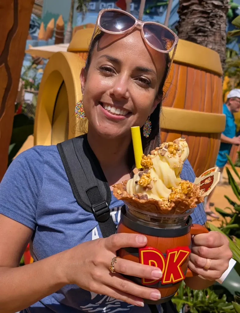 a Smiling Woman Holds a Dk-themed Mug Filled with Soft Serve Ice Cream and Caramel Popcorn at Super Nintendo World. She Wears Sunglasses on Her Head, Colorful Earrings, and a Blue Shirt, Standing Outdoors with Barrels and Palm Trees in the Background. - Favorite Grampy Travels