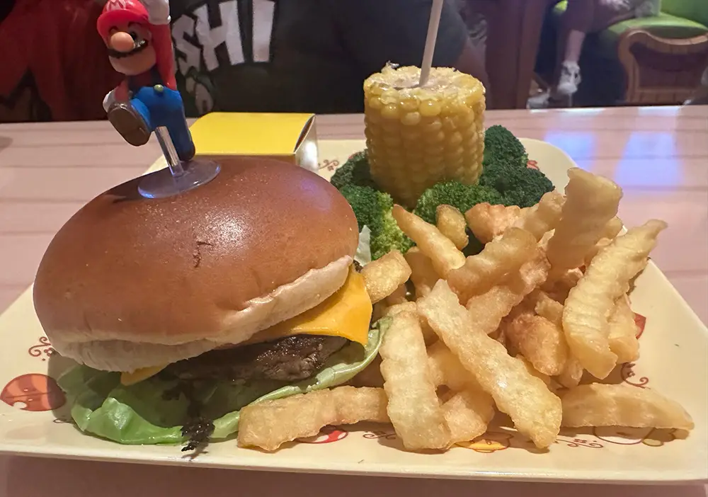 Cheeseburger with Lettuce and Cheese on a Bun, Topped with a Toy Figure on a Toothpick, Served with French Fries, Broccoli, and Corn on the Cob on a Plate.