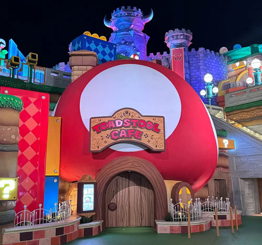 a Whimsical, Mushroom-shaped Building Labeled Toadstool Café Sits in a Colorful, Cartoon-like Setting Inspired by Super Mario, with Bright Blocks, Question Marks, and a Castle Visible in the Background. - Favorite Grampy Travels