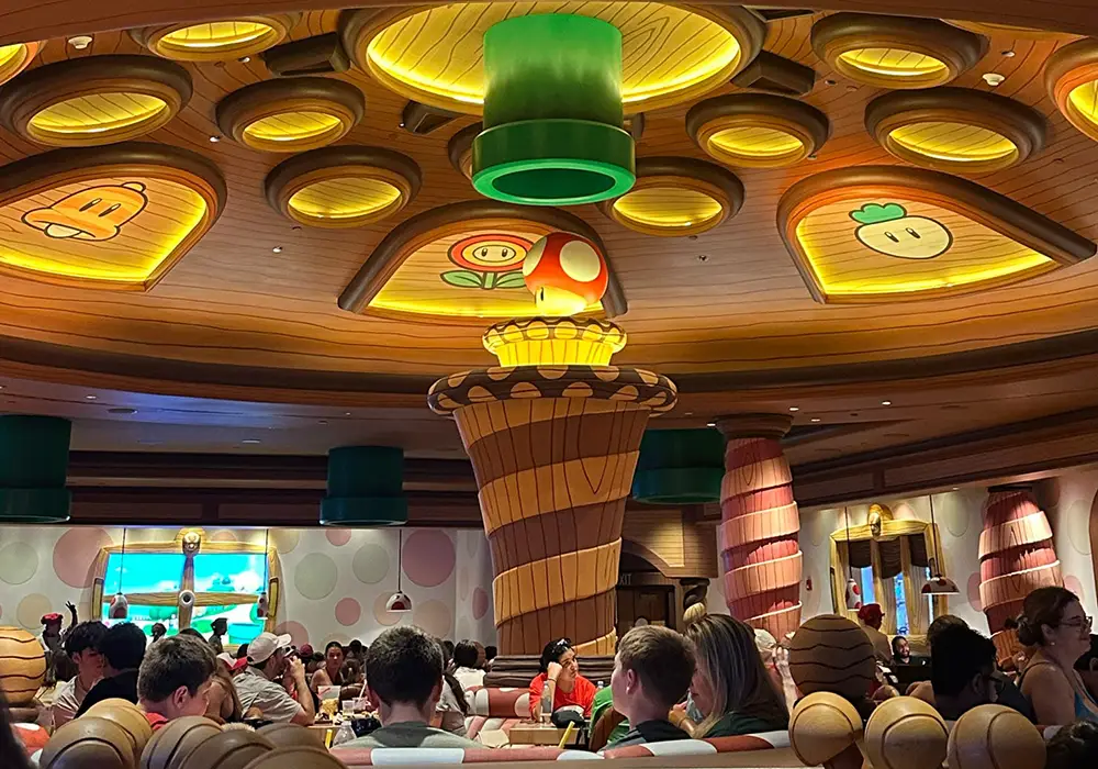 a Lively Restaurant with a Whimsical Ceiling Decorated with Glowing Cartoon-style Mushrooms and Power-ups, Inspired by Super Nintendo World. Perfect for Family Dining, Diners Sit at Round Tables Amid Colorful, Playful Video Game-themed Decor. - Favorite Grampy Travels