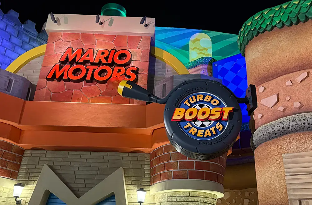 a Colorful Building Facade at Epic Universe Features a Sign Reading Mario Motors Above an Oversized Black and Yellow Lever Labeled Turbo Boost Treats. the Playful Design Has Brick and Stone Textures, Vibrant Lighting, and Classic Super Nintendo World Charm. - Favorite Grampy Travels