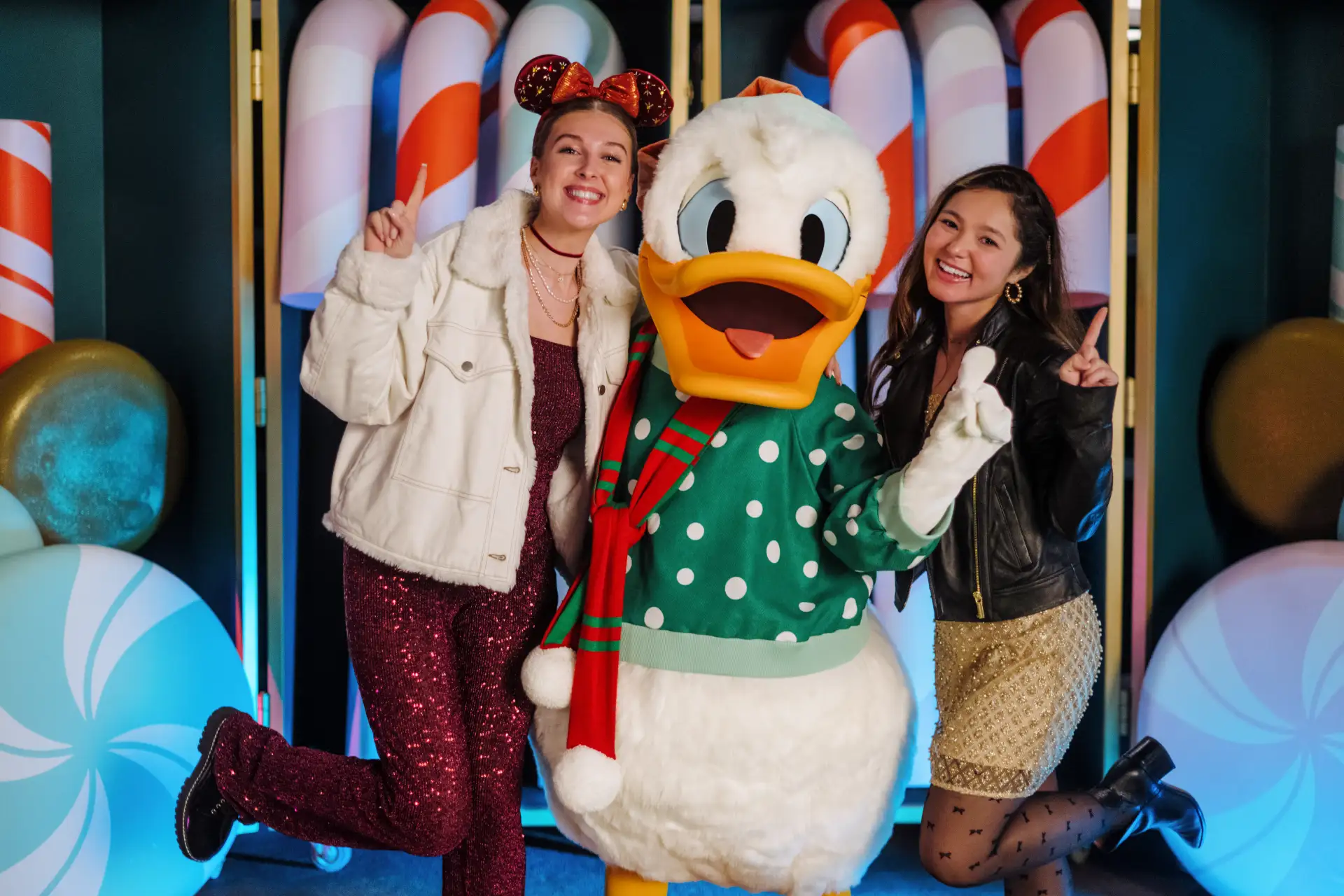 Two Smiling Women Pose with Donald Duck, Who is Dressed in a Green Polka-dot Sweater and Red Scarf, in Front of Large Christmas Candy Cane and Peppermint Decorations. All Three Are Making Playful, Cheerful Gestures at Disney's Jollywood Nights in Hollywood Studios in Walt Disney World - Favorite Grampy Travels - Favorite Grampy Travels 2026 - Favorite Grampy Travels 2026