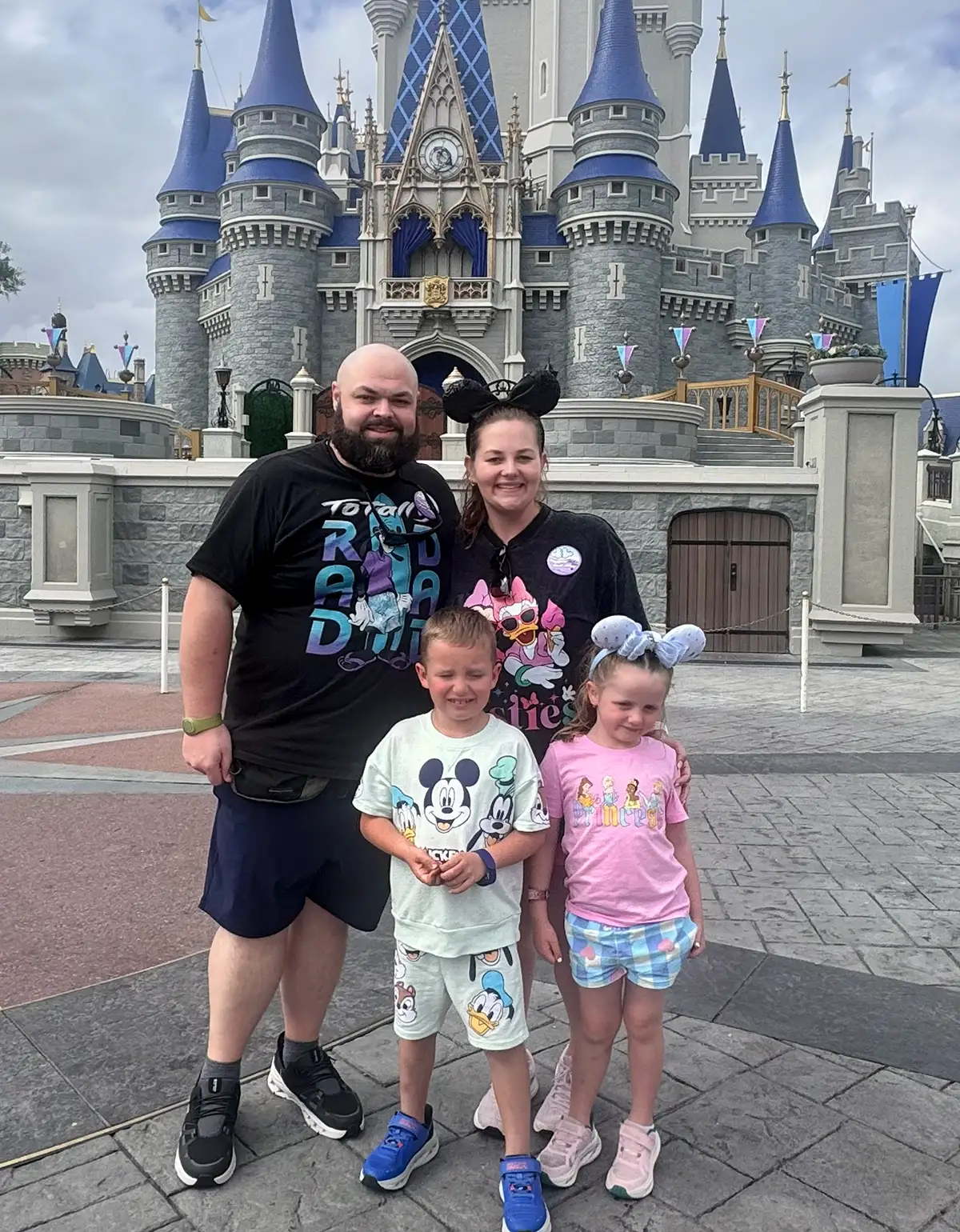 Samantha Malone and her smiling family of four pose in front of Cinderella Castle at a Disney park. The adults and children wear Disney-themed shirts and ears, with the castle’s blue and gray towers visible in the background. - Favorite Grampy Travels