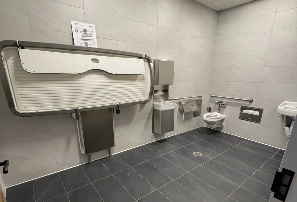 Accessible Public Restroom at Universal Kids Resort with Gray Tile Walls and Floor, Featuring a Large Adult Changing Table, Wall-mounted Toilet with Grab Bars, Paper Towel Dispenser, Trash Bin, and a Small Sink on the Right. - Favorite Grampy Travels - Favorite Grampy Travels 2026