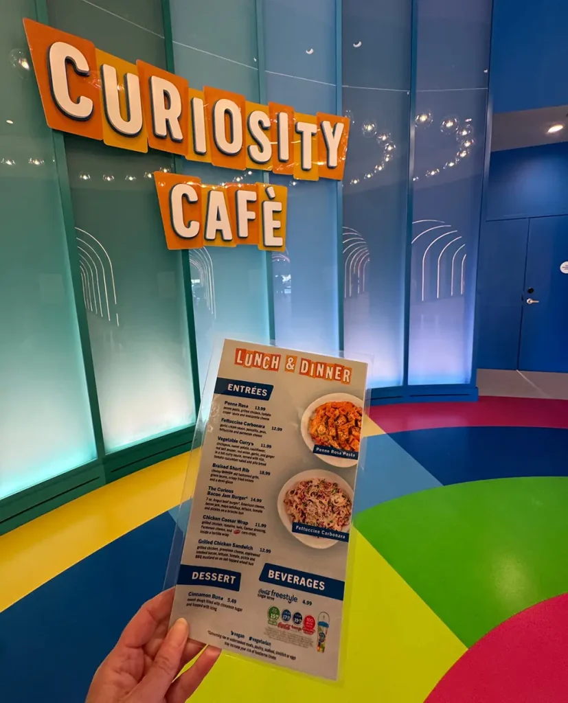 Ukr-curiosity-cafe-sign-and-menu - Favorite Grampy Travels a Hand Holds a Lunch and Dinner Menu in Front of a Colorful Wall with Large Orange Letters Spelling Curiosity Cafe. the Floor Features a Rainbow Pattern, and There is a Blue Door on the Right. - Favorite Grampy Travels - Favorite Grampy Travels 2026