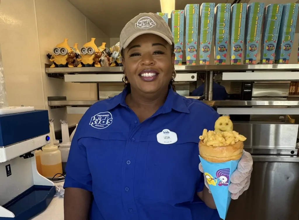 a Smiling Employee in a Blue Uniform and Tan Cap Holds a Food Cone Filled with Fries and a Character-shaped Item Inside Universal Kids Resort, with Minion-themed Food Containers on the Shelf Behind Her. - Favorite Grampy Travels - Favorite Grampy Travels 2026