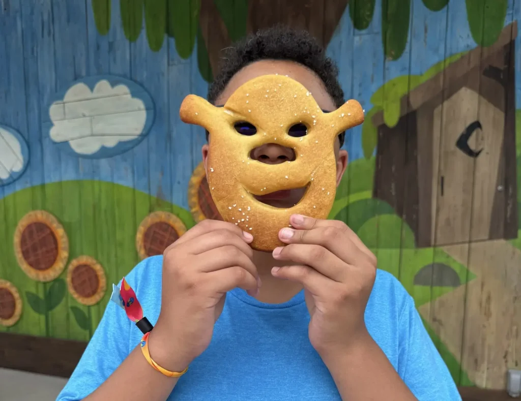 A child in a blue shirt holds a Shrek-shaped food cookie mask in front of their face, standing against a colorful, cartoon-style outdoor mural at Universal Kids Resort with trees, clouds, and sunflowers. - Favorite Grampy Travels