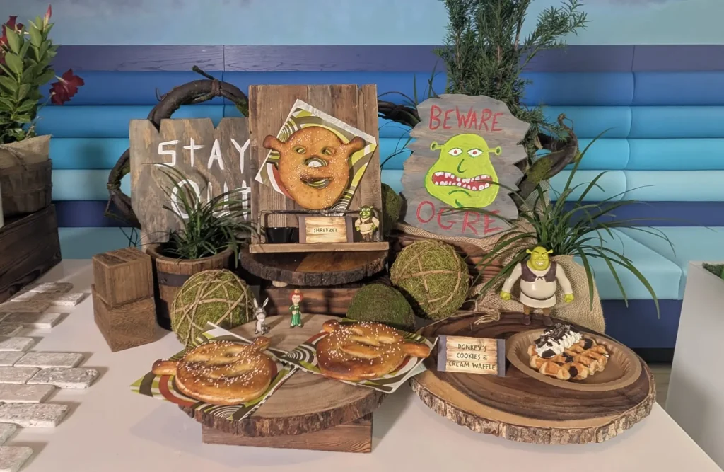 Display of Shrek-themed Bakery Props with Donuts on Wooden Platters and Signs Reading  - Favorite Grampy Travels 2026'BEWARE OGRE' and 'SHREKZEL' against a blue striped backdrop.