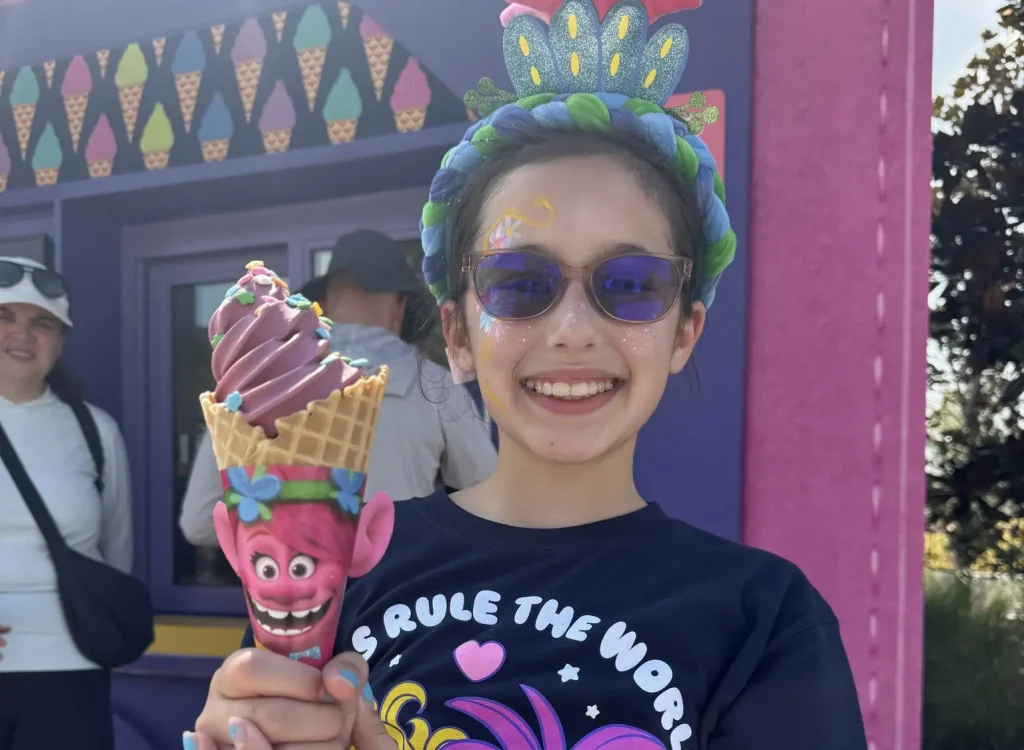Smiling Girl Wearing a Colorful Headband and Sunglasses Holds a Pink, Topped Ice Cream Cone with a Trolls Face on the Wrapper in a Bright Outdoor Setting. - Favorite Grampy Travels 2026