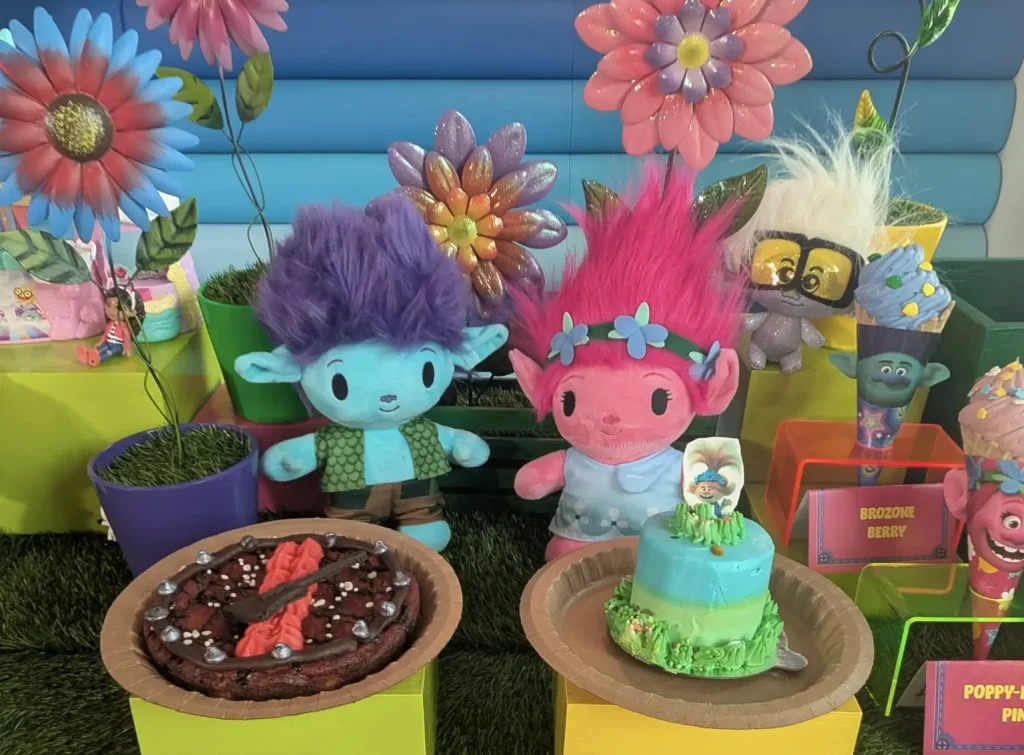 Stuffed Trolls Characters with Colorful Hair Are Displayed at a Party Table at Universal Kids Resort, Featuring a Blue and Pink Cake, Delicious Food, Chocolate Desserts, Large Artificial Flowers, and Brightly Colored Decor. - Favorite Grampy Travels - Favorite Grampy Travels 2026