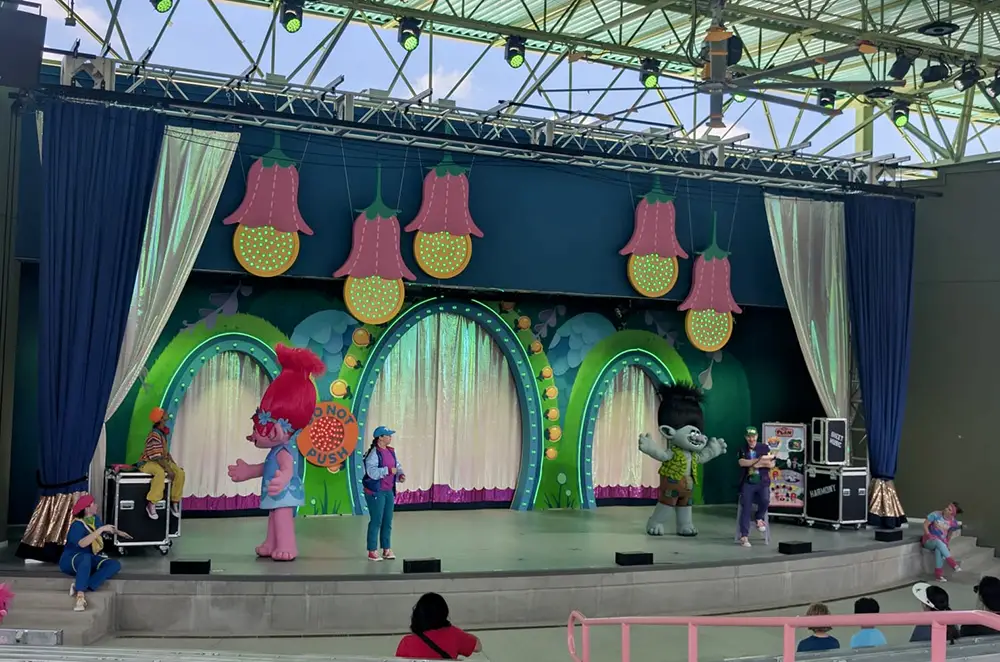 Costumed Characters, Including a Pink and Gray Troll, Perform on a Colorful, Flower-themed Stage with Bright Lights and Decorations. Several People Watch from the Audience in an Outdoor Amphitheater. - Favorite Grampy Travels - Favorite Grampy Travels 2026