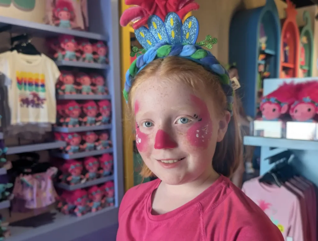 a Young Girl with Red Hair and Face Paint, Wearing a Colorful Headband with Flowers and Leaves, Smiles Inside a Store Filled with Bright Clothes and Plush Toys. - Favorite Grampy Travels - Favorite Grampy Travels 2026