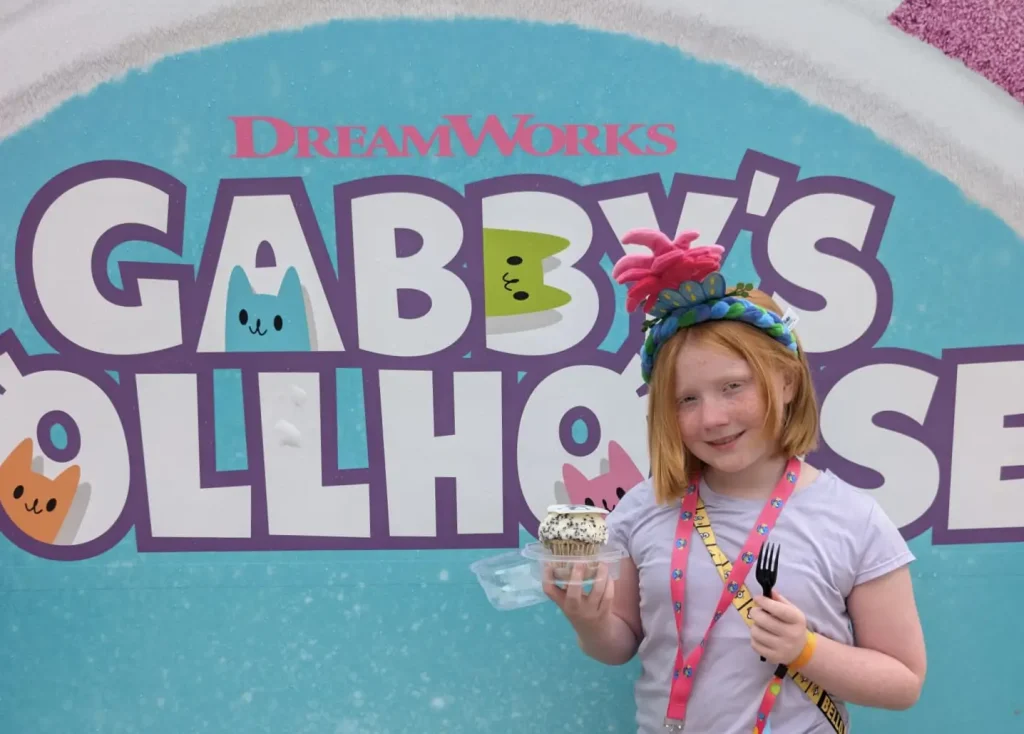 a Young Girl with Red Hair, Wearing a Colorful Flower Headband and Holding Delicious Food—a Cupcake with a Fork—stands Smiling in Front of a Gabby’s Dollhouse Dreamworks Sign with Cartoon Cat Faces at Universal Kids Resort. - Favorite Grampy Travels - Favorite Grampy Travels 2026