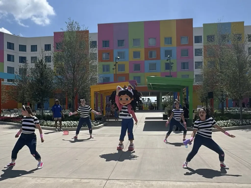 a Person in a Colorful Cartoon Cat Costume Stands Outside a Brightly Colored Building, Surrounded by Five Dancers in Matching Striped Shirts and Jeans, Performing Under a Sunny Sky. - Favorite Grampy Travels - Favorite Grampy Travels 2026