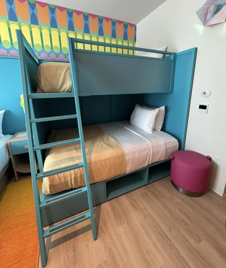 a Modern Room with Teal Bunk Beds; the Lower Bed is Neatly Made with White Sheets and a Tan Blanket. There’s a Colorful Patterned Wall, a Magenta Ottoman, and Wood Flooring. the Upper Bunk Has a Ladder for Access. - Favorite Grampy Travels - Favorite Grampy Travels 2026