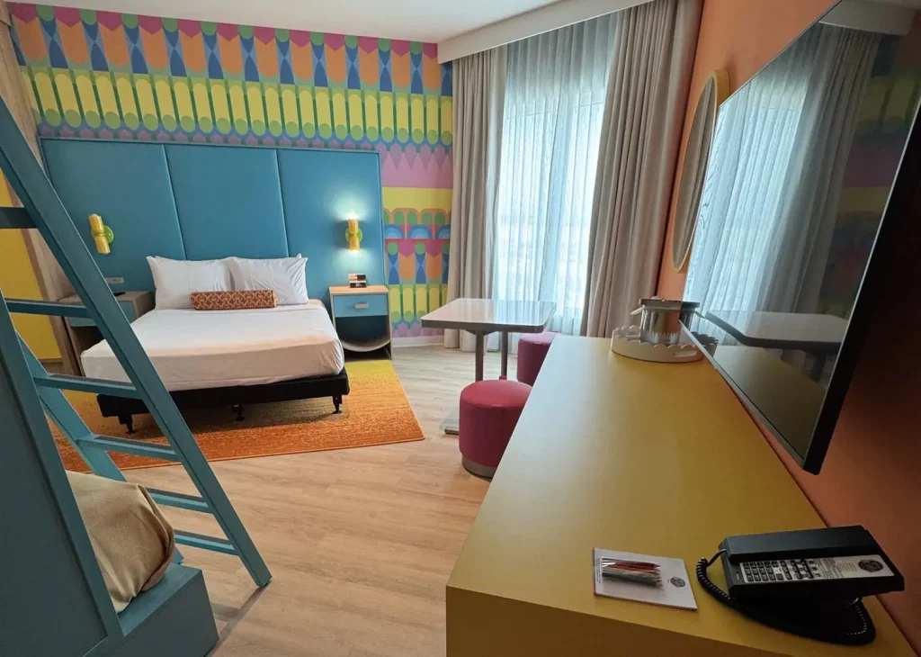 a Colorful Hotel Room at Universal Kids Resort Features a Double Bed, Blue and Orange Accent Wall, Bunk Bed Ladder, Round Table with Pink Stools, Large Windows with Sheer Curtains, Flat-screen Tv, and a Yellow Desk with a Phone. - Favorite Grampy Travels - Favorite Grampy Travels 2026