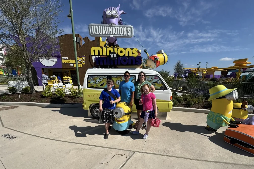 a Family of Four Poses and Smiles in Front of a Minions-themed Van and Characters at an Outdoor Attraction, with a Minions Vs Minions Sign—perfect Inspiration for Your Universal Kids Resort Planning. - Favorite Grampy Travels - Favorite Grampy Travels 2026