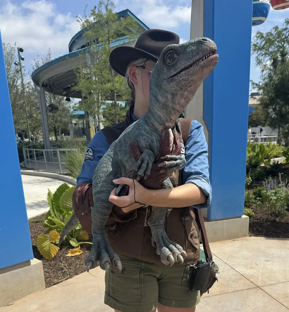 a Person in a Hat and Vest Holds a Large, Realistic Dinosaur Puppet Outdoors at a Theme Park, Surrounded by Greenery and Blue Structures. - Favorite Grampy Travels - Favorite Grampy Travels 2026