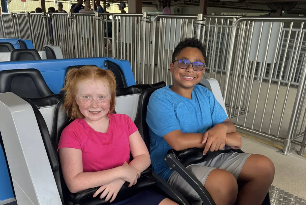 a Smiling Girl in a Pink Shirt and a Smiling Boy in a Blue Shirt Sit Side by Side in a Roller Coaster Car, Waiting for the Ride to Start. Metal Railings and the Loading Platform Are Visible in the Background. - Favorite Grampy Travels - Favorite Grampy Travels 2026