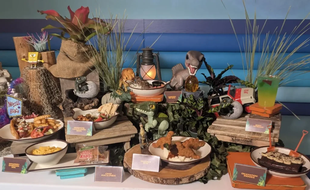 a Display of Dinosaur-themed Food and Drinks on Wooden Boards, Surrounded by Toy Dinosaurs, Leaves, and Props Like a Lantern and Toy Jeep—perfect for a Universal Kids Resort-inspired Party, All Set Against a Blue-striped Wall. - Favorite Grampy Travels - Favorite Grampy Travels 2026