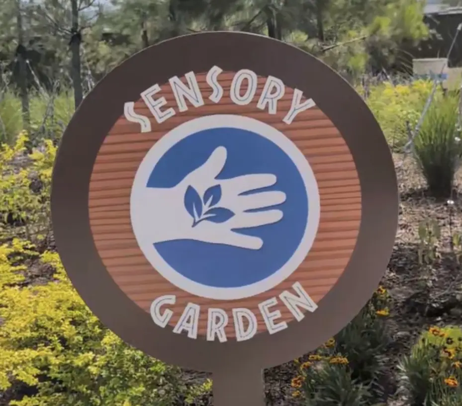 Ukr-jurassic-world-adventure-camp-sensory-garden - Favorite Grampy Travels Circular Sensory Garden Sign with a Hand Holding Leaves, Set Among Yellow Flowers and Greenery in Jurassic World Adventure Camp at Universal Kids Resort - Favorite Grampy Travels 2026