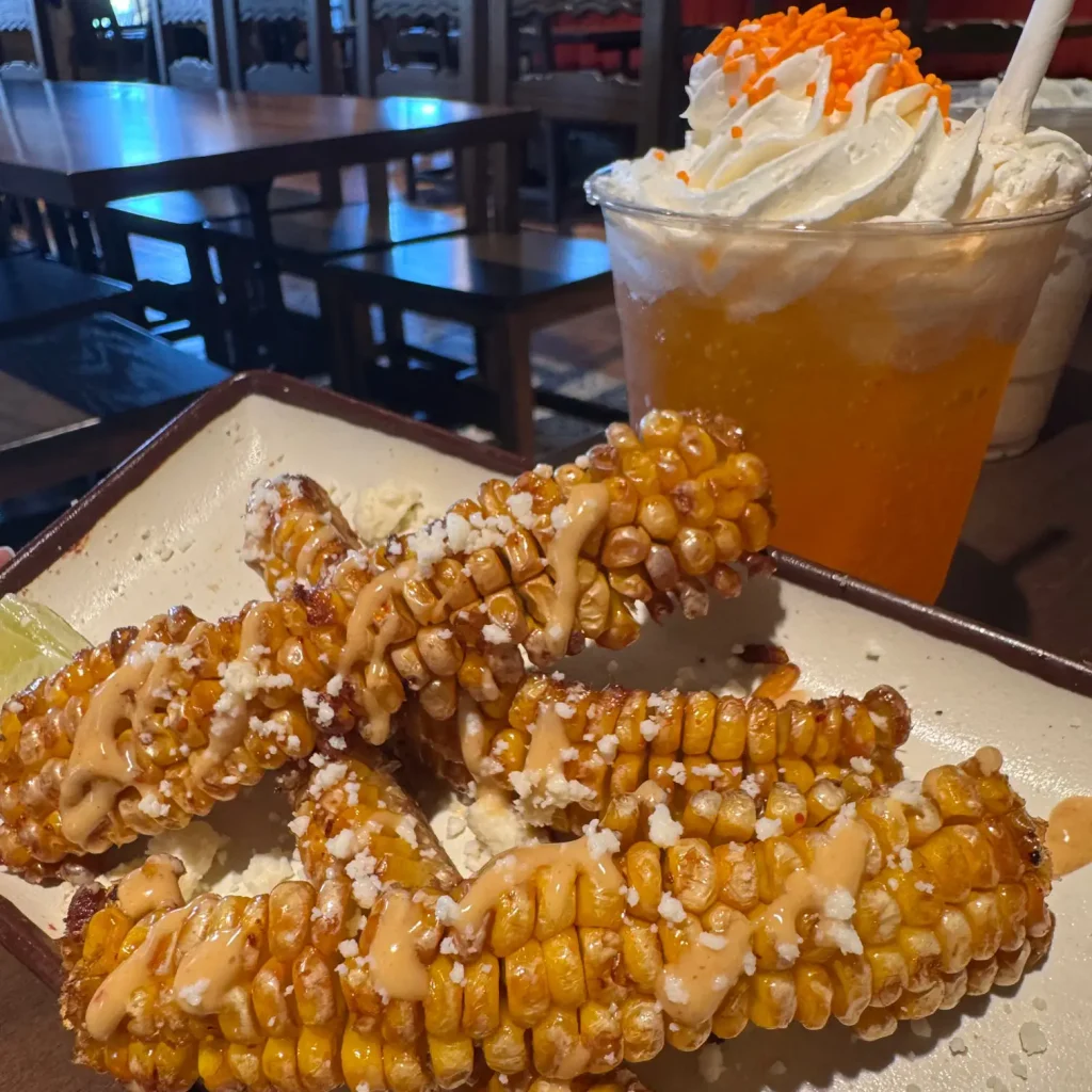 Corn on the Cob Skewers with Cheese Sauce and Crumbles on a Plate, with an Orange, Whipped-cream-topped Drink in the Background. - Favorite Grampy Travels 2026