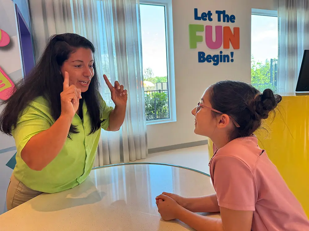 A woman in a green shirt enthusiastically talks to a smiling girl at a table. Colorful text on the wall reads, Let The FUN Begin! Family Services at Universal Kids Resort ensure every moment shines with cheerful decor and bright windows in the background. - Favorite Grampy Travels