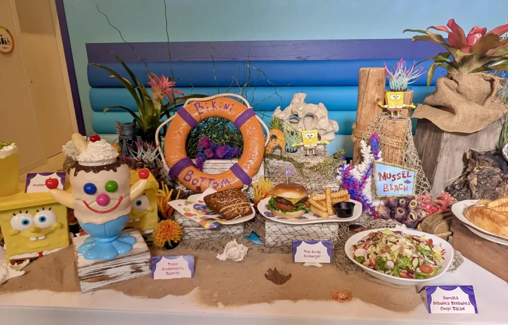 a Colorful Spongebob Squarepants-themed Food Display at Universal Kids Resort Features Character Figurines, a Bikini Bottom Lifebuoy, Themed Dishes Like Burgers, Salads, Desserts, All Set on Sand with Ocean-inspired Decorations and Labeled Cards. - Favorite Grampy Travels - Favorite Grampy Travels 2026