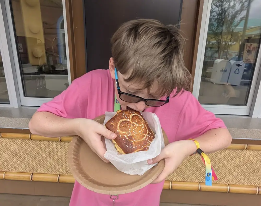 a Young Person Wearing Glasses and a Pink Shirt Bites into a Large Pretzel Shaped Like Mickey Mouse, Holding It on a Paper Plate Outdoors. - Favorite Grampy Travels - Favorite Grampy Travels 2026