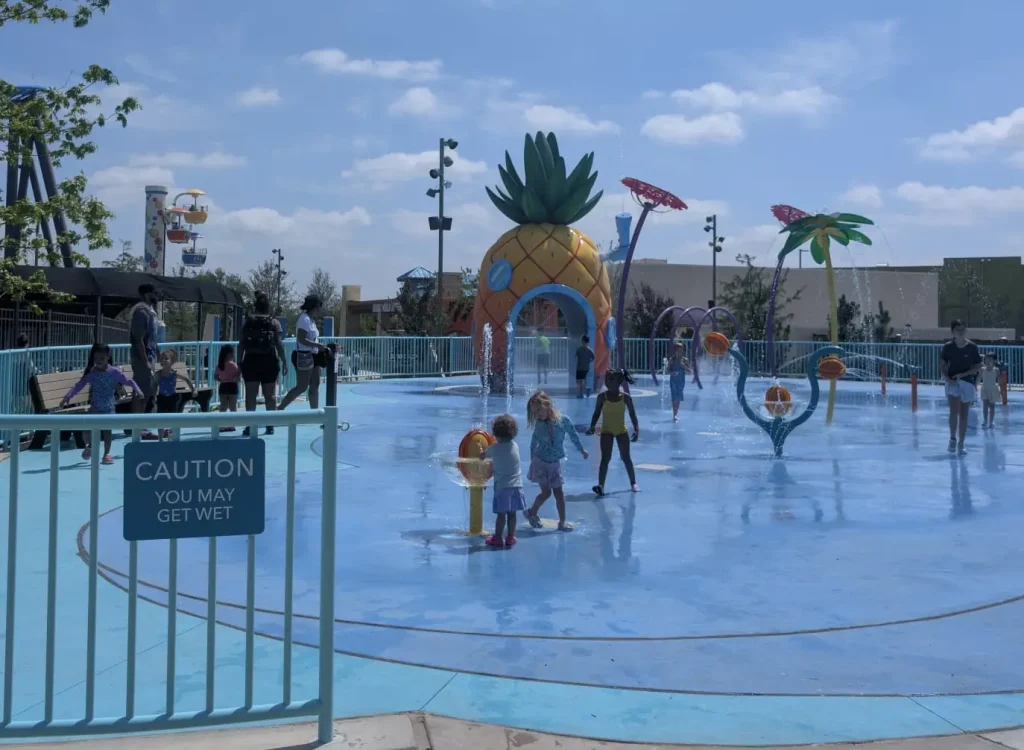Children and Adults Play at a Splash Pad with Water Features Shaped Like a Pineapple and Palm Trees. a Blue Sign Reads, Caution You May Get Wet. the Sky is Partly Cloudy. - Favorite Grampy Travels - Favorite Grampy Travels 2026