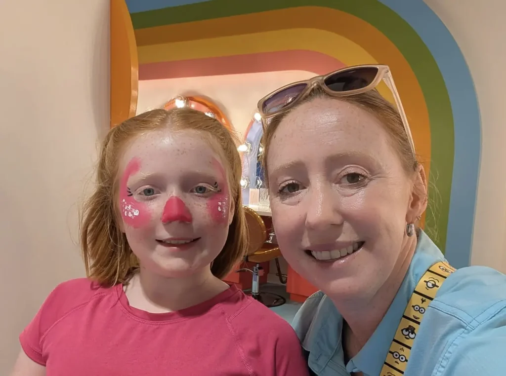 A child with pink face paint and a woman smiling pose for a selfie in front of a rainbow-colored wall at Universal Kids Resort. The woman wears sunglasses on her head and a Minions lanyard. Both look happy. - Favorite Grampy Travels