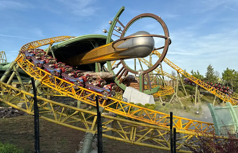 a Brightly Colored Roller Coaster with Yellow Tracks Winds Around a Large Metallic Sphere and Ring Structure at Epic Universe, with Riders Speeding Along Under a Blue Sky and Surrounding Greenery. - Favorite Grampy Travels - Favorite Grampy Travels 2026